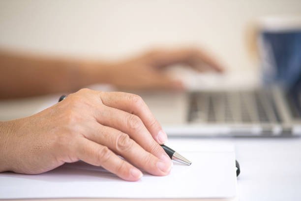 Close up: A womans hand holding a pen pointing at a notebook, the other hand on a laptop